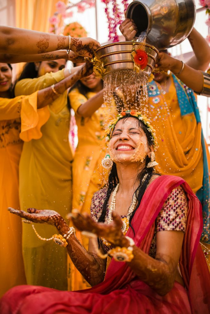 Smiling woman during a traditional Indian Haldi ceremony surrounded by friends pouring water in celebration. event planner